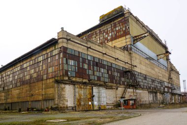 Paslı panelleri, kırık camları ve gri gökyüzünün altında eskimiş beton duvarları olan büyük terk edilmiş bir fabrika.