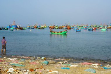 Mui Ne, Vietnam- 22 Mart 2025: Plastik atıklarla dolu bir kıyıda tek başına işeyen bir adam renkli balıkçı teknelerini izliyor.