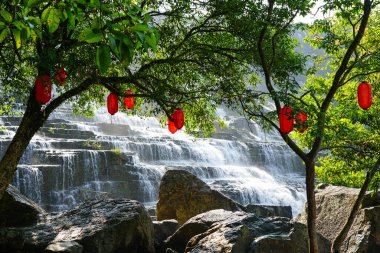 Yükselen Pongour Şelalesi yeşil ağaçlardan sarkan canlı kırmızı fenerlerin arkasında akmakta, doğa ve kültürü harmanlayarak.