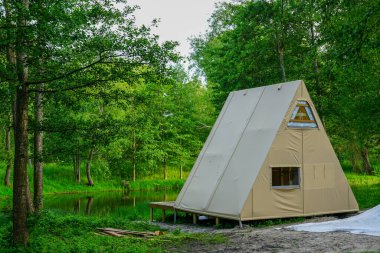 Yeni yapılmış bir A çerçeveli glamping kabin, görünür ahşap çerçeveli bej bir muşamba ile kaplı bir orman göleti yanında huzur içinde duruyor.