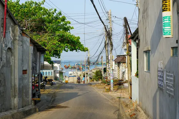 Mui Ne, Vietnam-March22, 2025: Dar, eğimli bir cadde, parlak bir sabah gökyüzü altında renkli balıkçı tekneleriyle dolu hareketli bir limana gider.