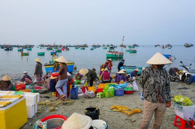 Mui Ne, Vietnam- 22 Mart 2025: Konik şapkalı yerli kadınlar sahilde taze deniz ürünlerini ayırırken renkli sepet botlar açık denizde yüzüyor