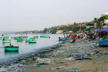 Mui Ne, Vietnam- 22 Mart 2025: Plastik atık yığınları işlek, kirlenmiş sahil pazarını kaplarken geleneksel balıkçı tekneleri kıyıda yüzüyor