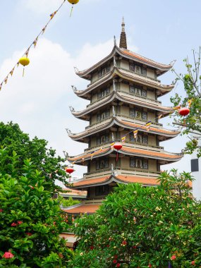 Vinh Nghiem Pagoda 'nın yüksek binası Ho Chi Minh şehrindeki canlı yaprakların üzerinde yükselir, fenerler ve dua bayraklarıyla süslenir.