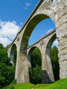 Polonya 'daki Stanczyki Köprüsü' nün yüksek taş kemerlerinin yoğun yeşil orman ve mavi gökyüzü ile çevrili yakın görüntüsü.