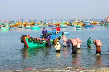 Mui Ne, Vietnam- 22 Mart 2025: Erkekler parlak renkli gemiler ve mavi sularla çevrili bir balıkçı teknesine ağır çuvallarla yürür.