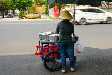Geleneksel konik şapka takan bir kadın, Vietnam 'da bir şehir caddesinde araçlar geçerken sokak arabasından yemek servisi yapıyor.