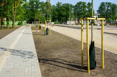 Ahşap çerçevelerle desteklenen genç ağaçlar, kentsel alanda yapım aşamasındaki bir yolun yanında yeni asfaltlanmış bir kaldırım hizasında.