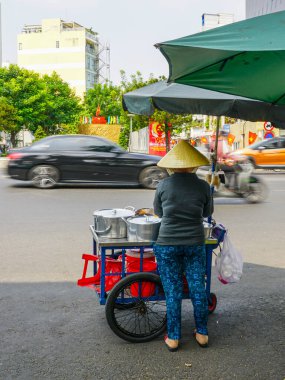 Konik şapkalı Vietnamlı bir kadın yeşil şemsiye altında yemek arabasını kullanırken kalabalık bir caddede trafik koşuşturuyor.