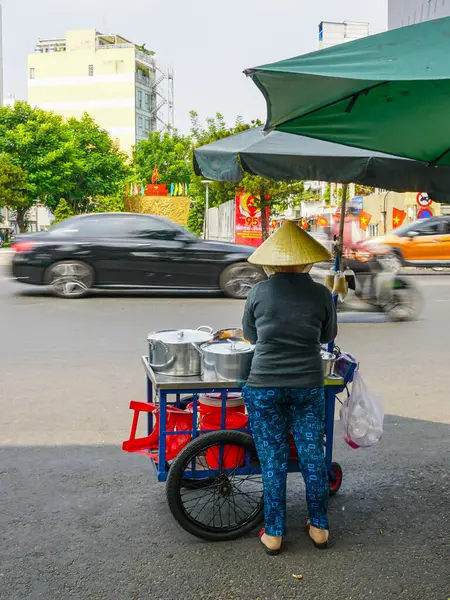 Konik şapkalı Vietnamlı bir kadın yeşil şemsiye altında yemek arabasını kullanırken kalabalık bir caddede trafik koşuşturuyor.