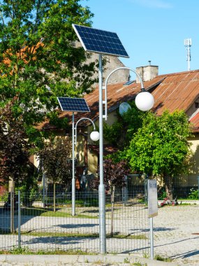 Ağaçları, çitleri ve mavi gökyüzünün altındaki paslı çatıları olan bir yerleşim bölgesinde enerji tasarruflu güneş ışığı.