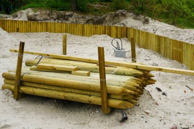 Arkasında ahşap istinat duvarı ve yeşillik olan kumlu bir şantiyeye istiflenmiş tahta direkler.