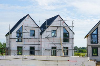 İki katlı modern bir ev, iskele, yalıtım panelleri, ve temiz mavi gökyüzünün altında tamamlanmamış dış duvarlar.