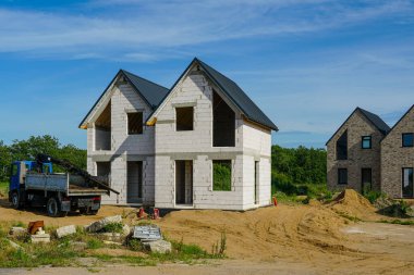İnşaat aşamasındaki modern iki katlı bir ev, açık mavi gökyüzünün altında bir kamyon ve inşaat malzemeleri var.