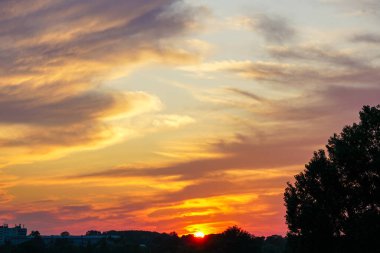 Canlı turuncu, sarı ve mor renkleriyle sersemletici bir gün batımı huzurlu siluetli bir manzara üzerinde dramatik bulutlar aydınlatıyor.