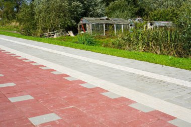 Yeni yapılmış kırmızı ve gri asfalt kaldırımlar, kırsal alanda yeni ve eski sembolize edilmiş eski, terk edilmiş seralarla tezat oluşturuyor.