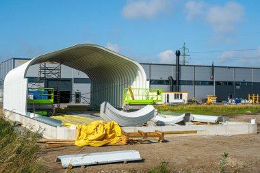 Prefabrik çelik yapı hangarı, kıvrımlı çatı panelleri, iskelesi ve bir sanayi bölgesinde ekipmanı var.