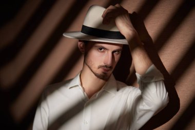 Zarif beyaz tişörtlü, beyaz şapkalı yakışıklı bir esmer adam karanlık ışıklı ve gölgeli bir odada duruyor. Erkek güzeli, moda. Kara film stili. 