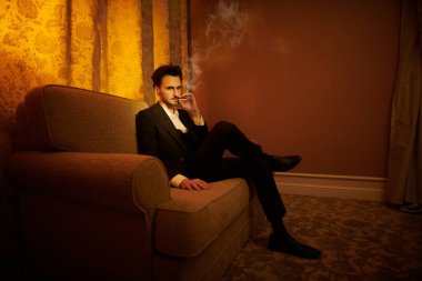 Lüks bir dairede kanepede oturup sigara içen yakışıklı bir esmer adamın portresi. Göz kamaştırıcı bir yaşam tarzı. Moda fotoğrafı.. 