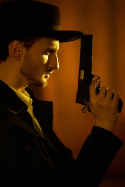 Siyah paltolu cesur bir esmer adam portresi ve elinde bir silahla, bir ışık huzmesi ile karanlık odada duran bir şapka. Retro noir tarzı. Suç dünyası, mafya..