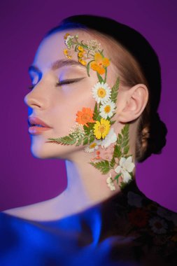 Yüzünde bahar çiçekleri olan güzel kızıl saçlı bir kız ışık çemberinin mor arka planında poz veriyor. Parfüm. Makyaj ve makyaj malzemeleri. Güzel atış.. 