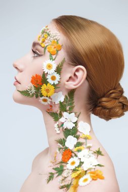 Yazın kokusu. Zarif makyajlı, yüzünde ve omzunda yaz çiçekleri olan çekici kızıl saçlı bir kız beyaz arka planda poz veriyor. Kozmetik ve parfüm. 