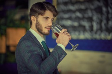 Zarif klasik takım elbiseli, kendine güvenen yakışıklı bir esmer adam lüks dairelerde dikilip bir bardak şarap içiyor. Erkek güzelliği, moda. İş stili. İşadamı. 