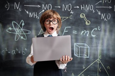 Okul üniformalı duygusal bir çocuğun portresi ve dizüstü bilgisayarı ile bilimsel formüller ve çığlıklarla karatahtanın önünde duran gözlük. Eğitim. 