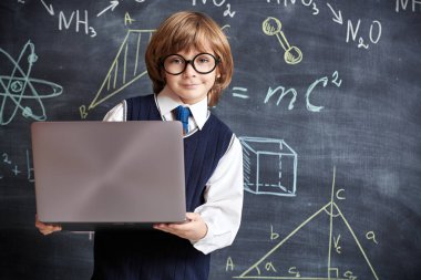 Okul üniformalı zeki bir çocuğun portresi ve arka planda bilimsel formülleri ve diyagramları olan bir dizüstü bilgisayar tutan gözlüklü. Akıllı çocuklar. Eğitim. 