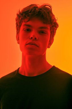 Dalgalı kahverengi saçlı yakışıklı huysuz bir adam turuncu arka planda kameraya kırmızı ışıkla bakıyor. Ergenler ve duygular. 