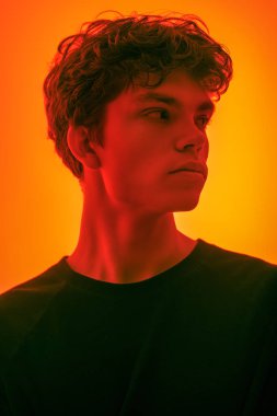 Kırmızı ışıklandırmalı, turuncu arka planda duran kapkara saçlı yakışıklı bir adamın stüdyo fotoğrafı. Gençler, duygular. 