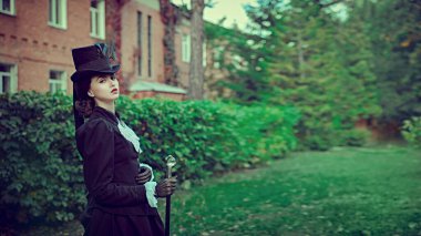 19. yüzyılın katı siyah takım elbiseli, kendinden emin ve sofistike bir bayan yeşil yapraklarla çevrili tuğla bir evde yürüyor. Boşluğu kopyala. 