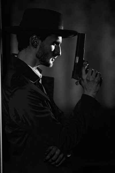 Black-and-white portrait of a courageous handsome man in black coat and a hat standing in a dark room, lit by a ray of light. Retro noir style. Criminal world, mafia.