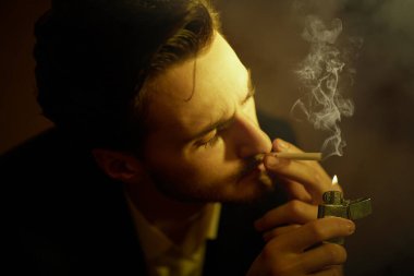 Zarif siyah takım elbiseli kendine güvenen yakışıklı bir esmer karanlık bir odada sigara yakıyor. Kara film stili. Erkek güzelliği, moda. 