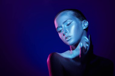 Studio shot of a graceful fashion model with shiny silver skin in colorful neon blue and pink light. Beauty and cosmetics concept. Body painting. Dark blue background with copy space. clipart