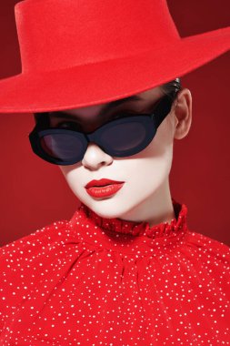 Kırmızı elbiseli, kırmızı şapkalı ve güneş gözlüklü zarif genç bir kadının moda fotoğrafı. Kırmızı arka plan. Güzellik, makyaj ve kozmetik. 