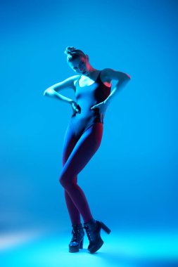 Stüdyoda siyah bir elbise, kırmızı tayt ve yüksek topuklu çizmelerle poz veren ince model bir kızın tam boy portresi. Mavi arka planda karışık mavi ve kırmızı ışık. Yüksek moda çekim. 