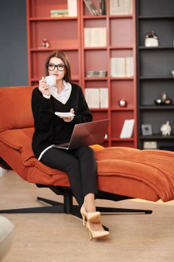 Modern bir ofiste bir fincan çayla kanepede oturan güzel, modern bir iş kadını. Modern iç mekan ve mobilya.