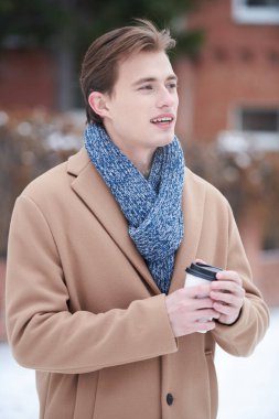 Havalı bir palto ve eşarp giymiş şık bir genç adam kış günü dışarıda durup kahve içiyor. Moda fotoğrafı. Erkek yaşam tarzı..