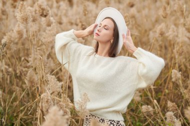 Beyaz yün kazaklı ve beyaz keçeli şapkalı zarif genç kadın sonbahar parkında sazlıklar arasında poz veriyor. Zarif sonbahar modası ve güzelliği. 