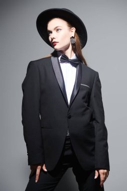 Mükemmel görünüş. Gri bir arka planda poz veren siyah takım elbiseli, papyonlu ve şapkalı güzel bir mankenin moda fotoğrafı. İş kıyafetleri tarzı. 