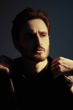 Deri ceketli yakışıklı, vahşi, esmer bir adam ışıkla gölge arasında karanlık bir zeminde dikiliyor. Erkek güzelliği, moda. Duygular.