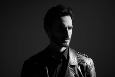 Siyah-beyaz bir esmer adam portresi şık bir deri ceket giymiş koyu bir arka planda duruyor ve yoğun bir şekilde yüzünü çeviriyor. Erkek güzeli, moda. Duygular.