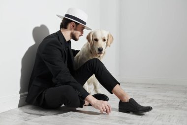 Siyah takım elbiseli, beyaz şapkalı yakışıklı, zarif, esmer bir adam Golden Retriever köpeğiyle beyaz bir duvara yaslanmış. Erkek modası. Tam boy stüdyo resmi..