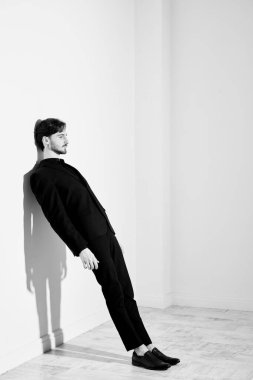 Siyah takım elbiseli esmer bir adamın beyaz bir duvara yaslanmış tam boy siyah beyaz portresi. Umutsuz durumlar. İnsanlar ve duygular.