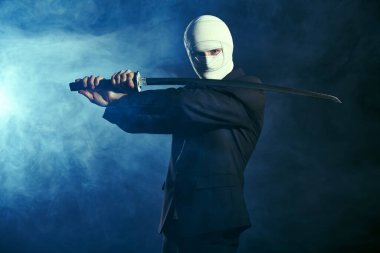 Siyah takım elbiseli savaşçı bir işadamı, sargılı bir yüzle, bir katana samuray kılıcıyla savaşa hazırdır. Rekabetçi savaşlar, iş kavramı. Dumanlı karanlık bir arkaplanda stüdyo portresi.