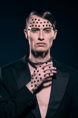 Moda fotoğrafı. Siyah kuyruklu yakışıklı, vahşi bir adamın portresi. Yüzünde ve elinde siyah bir arka planda metal çiviler var..