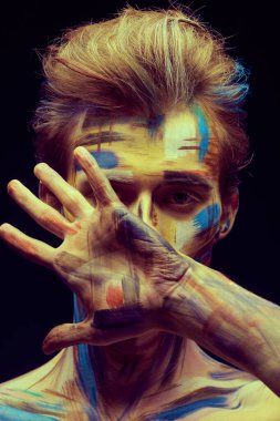 Vücut boyama projesi. Yüzünü eliyle kaplayan renkli boyanmış derili yakışıklı bir adamın portresi. Siyah arka plan. Sanat ve moda.