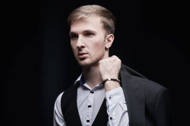 Erkek modası. Beyaz gömlekli ve yelekli yakışıklı bir adam koyu bir arka planda omzunda bir ceket tutuyor. Şık bir akşam stili.. 