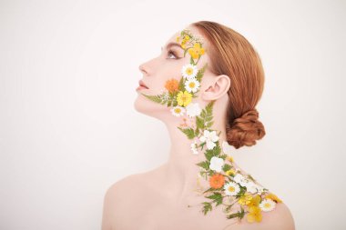 Güzel, kızıl saçlı, zarif makyajlı ve yüzünde yaz çiçekleri olan genç bir kadın. Beyaz arka plan. Parfüm. Makyaj ve makyaj malzemeleri. Güzellik ve çiçekler.